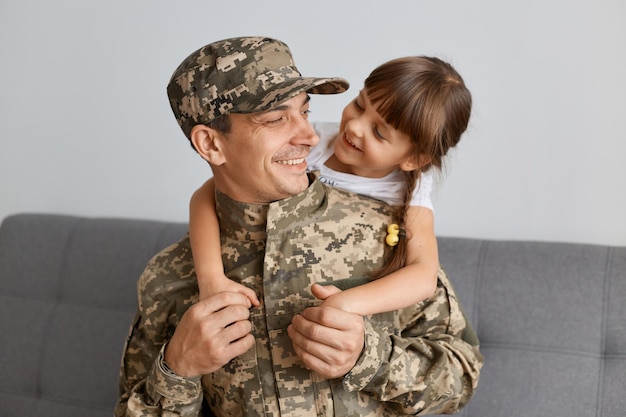 Image of happy delighted man soldier wearing camouflage uniform
and cap posing with his daughter hugging each other male came back
home after war or army