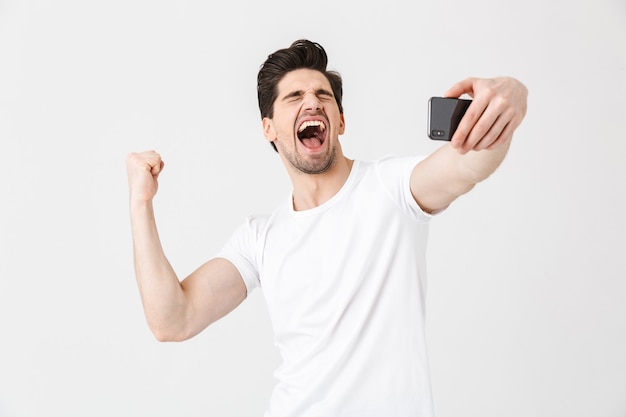 L'immagine di un giovane emotivo felice in posa isolato su un muro bianco prende selfie con il telefono cellulare che mostra il gesto del vincitore.