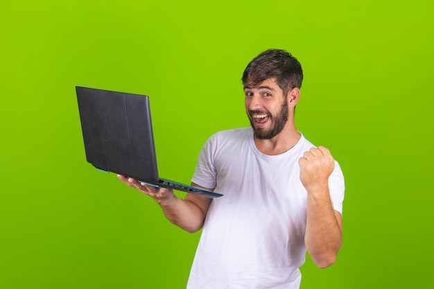 ラップトップ コンピューターを使用して緑の壁を越えてポーズをとって幸せな興奮した若い男のイメージは、勝者のジェスチャーを作る