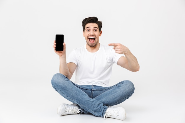 바닥을 가리키는 디스플레이에 앉아 있는 휴대폰을 사용하여 흰 벽 위에 고립된 채 포즈를 취하는 행복한 흥분한 젊은 남자의 이미지.
