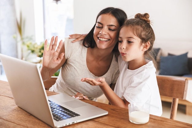 Image of happy family woman and her little daughter smiling and waving at laptop computer together while sitting at table in apartment