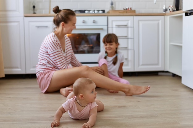 Image of happy family woman wearing striped casual style shirt sitting on floor in kitchen with daughters toddler baby crawling mother talking with elder kid