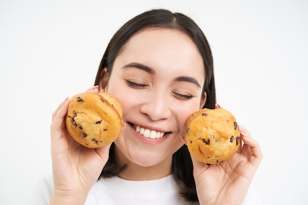 Image of happy korean girl who loves pastry smiling and eating cupcakes isolated on white background