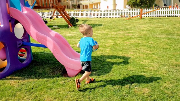 Image of happy laughing cheerful boy holding football ball in hands and running on children playground at park