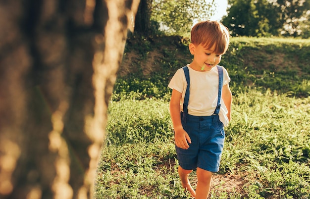Image of happy little boy wearing blue shorts smiling and playing at sunlight and nature background Adorable child running on the green grass in the park Kid having fun Childhood
