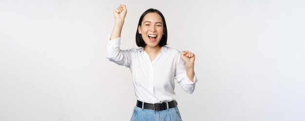 Image of happy lucky asian woman hooray gesture winning and celebrating triumphing raising hadns up and laughing standing over white background