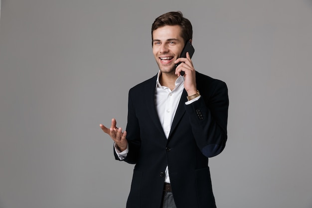 Image of happy man 30s in business suit talking on black smartphone, isolated over gray wall