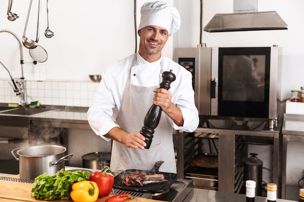 Image of happy man chief in white uniform cooking meal with meat and vegetables, at kitchen in restaurant