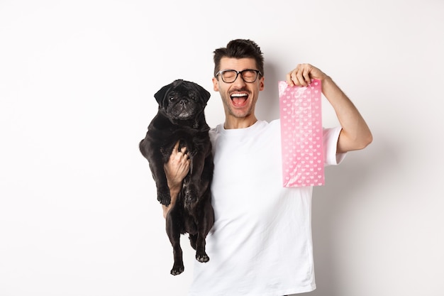 Image of happy man, pet owner, holding cute black pug and dog poop bag, standing over white