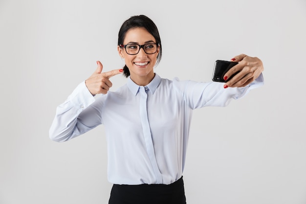 白い壁に隔離された携帯電話でselfie写真を撮る眼鏡をかけている幸せなオフィスの女性の画像