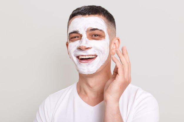 Image of happy optimistic man wearing white t shirt doing skin care procedures with pleasure looking at camera with smile posing isolated over gray background