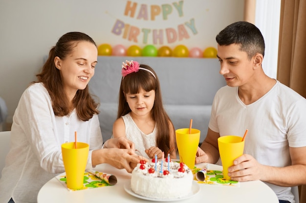 Image of happy positive family celebrating good event sitting at table with birthday cake and drink charming kid cutting holiday dessert together with mother expressing good emotions