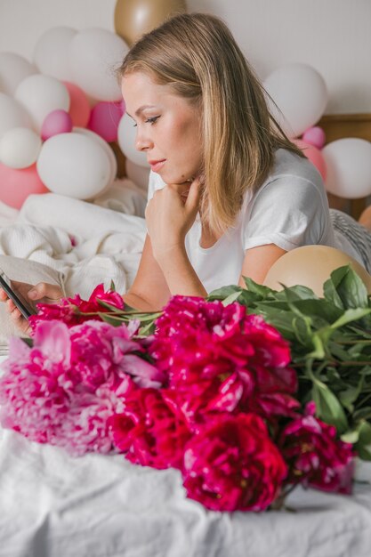 Image of a happy positive pretty young woman indoors at home  in bedroom in bed holding flowers take a selfie by mobile phone.