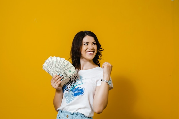 Изображение счастливой довольной девушки, держащей кучу денежных банкнот и выставочных жестов, изолированных на желтой стене
