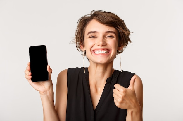 Image of happy satisfied woman recommending something, showing thumbs-up and smartphone screen, standing over white.