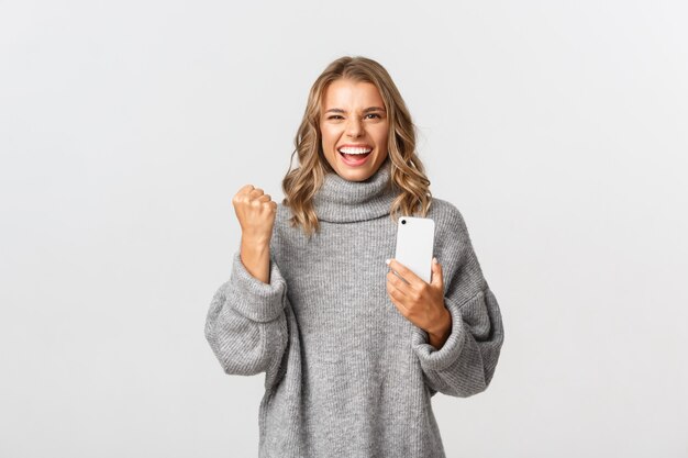 Image of happy winning girl in grey sweater, saying yes and triumphing, holding mobile phone