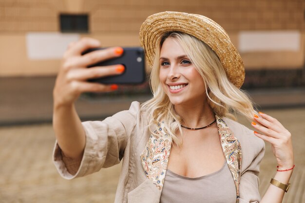 Image of a happy young cute woman walking outdoors take a selfie by mobile phone.