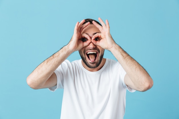 Image of happy young funny bearded man posing isolated over blue wall  showing glasses gesture with hands having fun showing tongue.