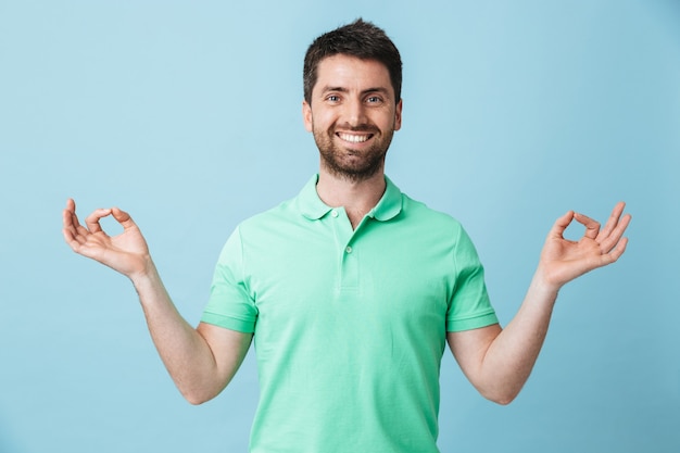 Image of a happy young handsome bearded man posing isolated over blue wall showing okay gesture meditate.