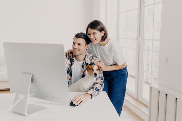 Image of happy young husband and wife focused in modern computer make shopping or watch video together spend time at apartment in living room pedigree dog poses at males knees collaborate