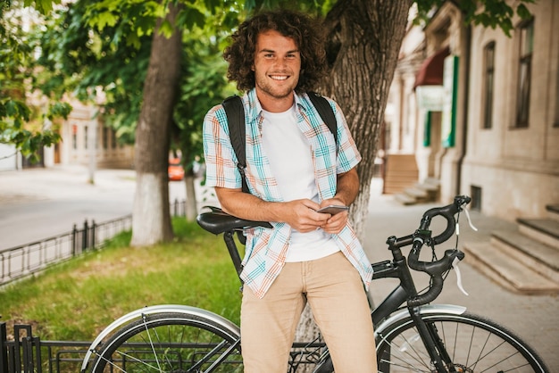 Image of happy young male student smiling with curly hair wearing shirt with backpack sitting on the bicycle relaxing outdoors browsing websites while shopping online on smart phone