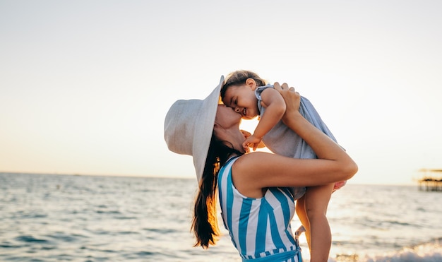 Image of happy young mother playing with her daughter at the sea sunset Beautiful female kissing baby walking outside at the ocean beach on sunset Motherhood love care