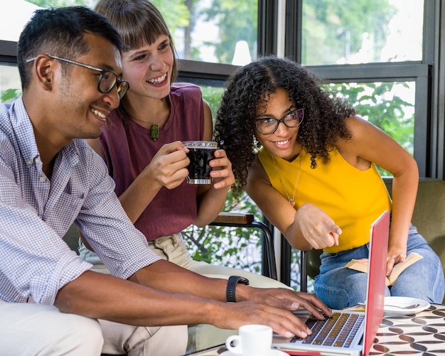 Изображение счастливых молодых людей, использующих ноутбук, сидя в кафе, молодой афроамериканки, указывающей устройство, многонациональной группы студентов в кафе, работающих на ноутбуке