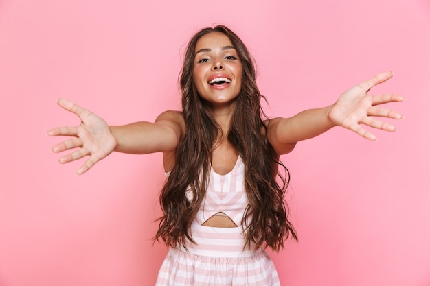 Image of happy young woman 20s with long hair wearing dress laughing and reaching arms towards you, isolated over pink wall