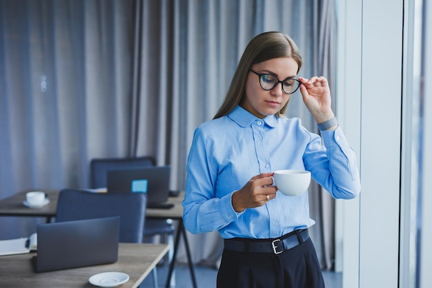 Immagine di una giovane donna felice con una camicia classica e occhiali che sorride e beve caffè mentre è in piedi vicino alla finestra in un ufficio moderno con grandi finestre lavoro a distanza