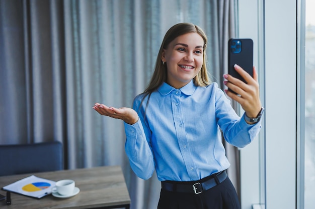 Изображение счастливой молодой женщины в куртке, улыбающейся и работающей на ноутбуке, разговаривающей по телефону в современном офисе с большими окнами Удаленная работа