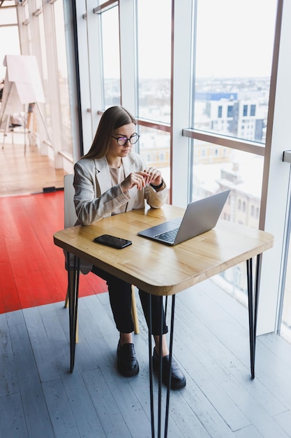 Изображение счастливой молодой женщины в куртке, улыбающейся и работающей на ноутбуке во время разговора по телефону в современном офисе с большими окнами Удаленная работа