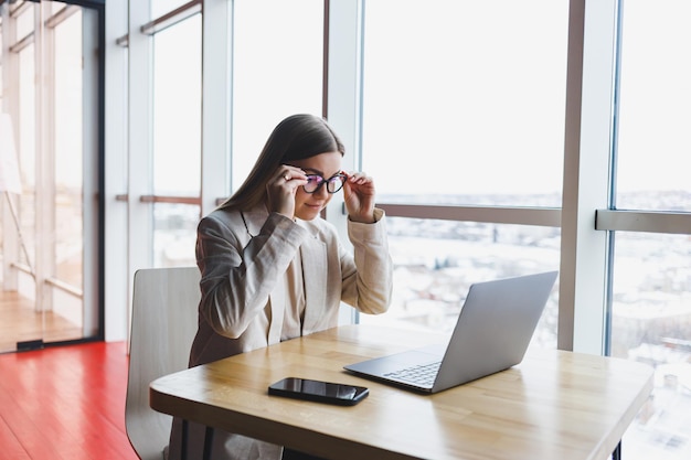 Immagine di una giovane donna felice con una giacca che sorride e lavora su un laptop mentre parla al telefono in un ufficio moderno con grandi finestre lavoro a distanza