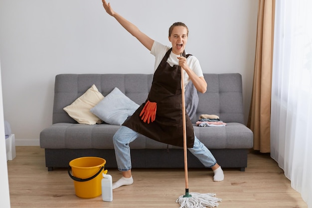 Image of happy young woman singing while washing floor in living room using wet moist mop as microphone having fun during house chores enjoying domestic duties gamifying cleaning routine