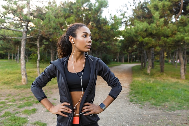 Image of healthy woman 20s wearing black tracksuit and earphones working out, while running through green park