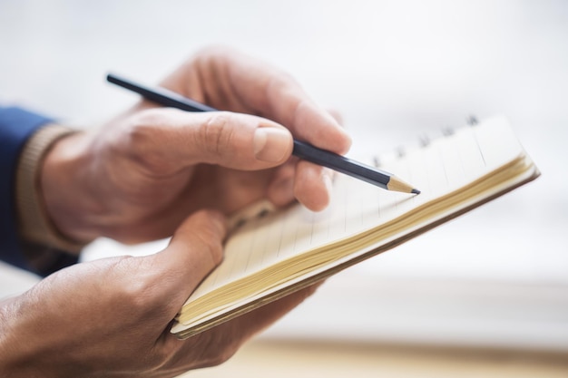The image highlights a man's hand writing in a notepad with a faintly blurred surrounding