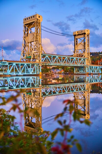 Image of Houghton sunrise of soft blues and purples and the lift bridge reflected in mirror like river of water and a blurred plant nearby