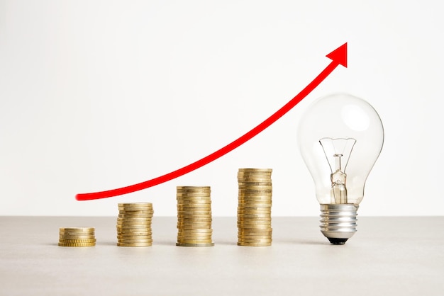 Image of increasing stacked coins a transparent light bulb with a rising red arrow above it on a neutral background