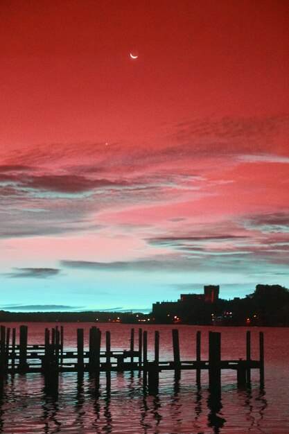 Инфракрасное изображение силуэта пустых доков в пурпурно-бирюзовой реке с красно-пурпурным небом с полумесяцем и небесно-голубыми облаками на восходе солнца