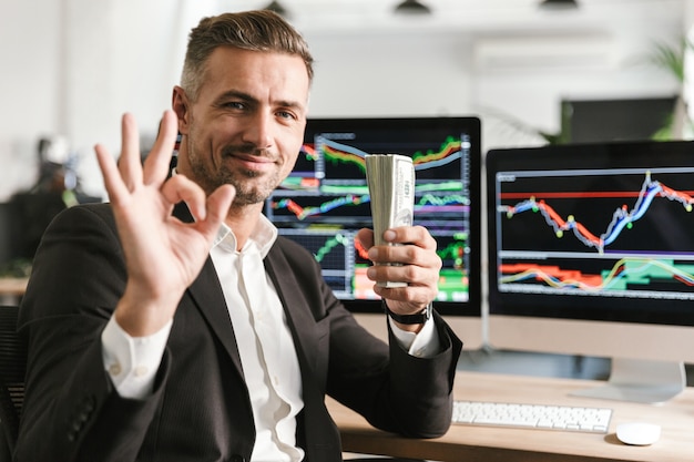 Image of joyful businessman 30s wearing suit holding pack of money while working in office with graphics and charts on computer