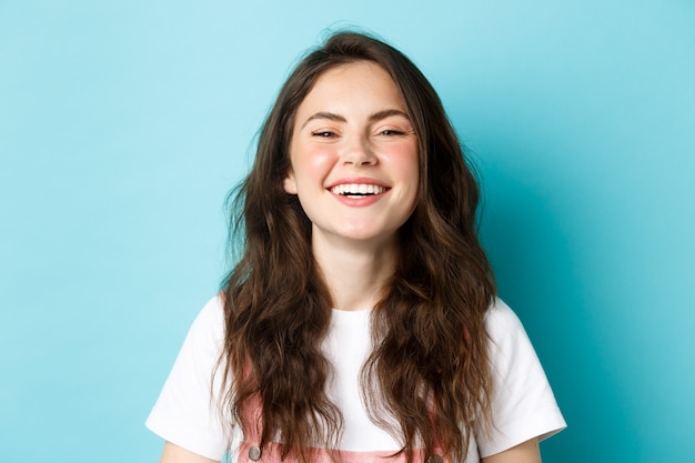 Image of joyful caucasian girl having fun, smiling with teeth, laughing over something funny, standing against blue wall