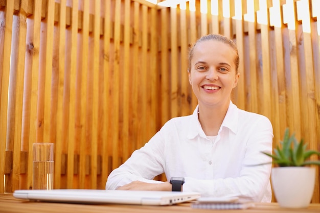 Image of joyful cheerful dark haired woman with bun hairstyle wearing white shirt sitting looking at camera with happy facial expression finishing her work on laptop