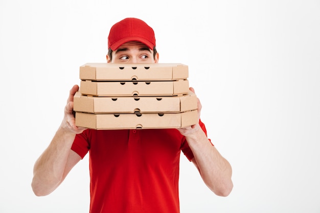 Image of joyful delivery man in red uniform covering face with stack of pizza boxes and looking aside, isolated over white space