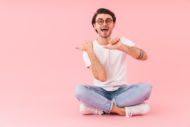 Image of joyful man wearing eyeglasses pointing fingers at copyspace while sitting with legs crossed isolated