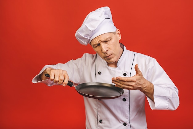 Immagine dell'uomo senior senior allegro in uniforme del cuoco che sorride e che tiene padella isolata sopra il fondo rosso della parete.