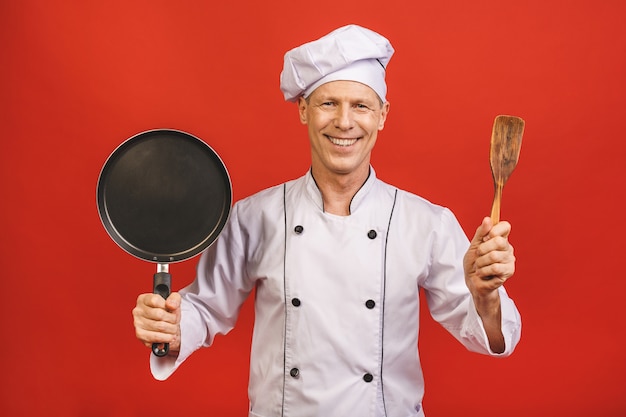 Изображение радостного старшего главного человека в форме повара усмехаясь и держа сковороду изолированный над красной предпосылкой стены.