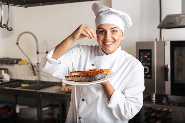 Image of joyful woman chef wearing white uniform, holding plate with grilled fish in kitchen at the restaurant