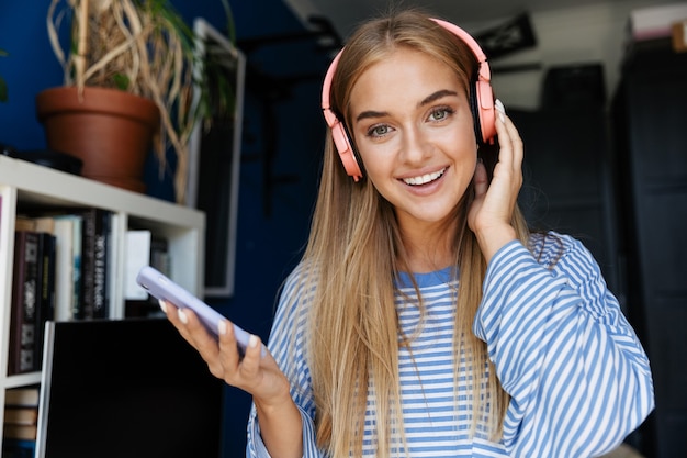 Image of joyful young girl wearing striped shirt using cellphone and headphones in apartment