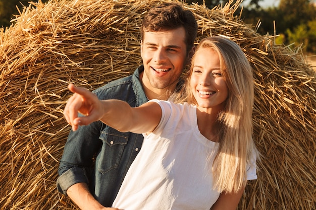 Image of joyous couple man and woman walking on golden field after harvesting, and standing near big haystack