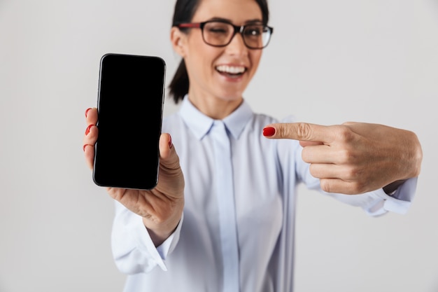 Immagine della donna gioiosa dell'ufficio che indossa occhiali da vista che tengono il telefono cellulare, isolato sopra il muro bianco