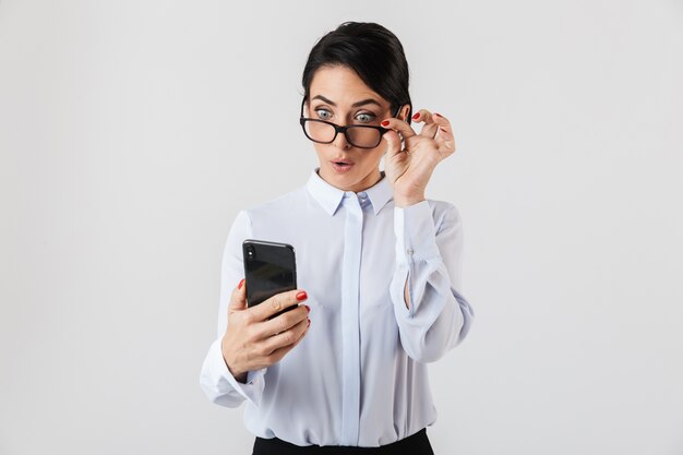 Immagine di gioiosa segretaria donna che indossa occhiali da vista utilizza lo smartphone in ufficio, isolato sopra il muro bianco